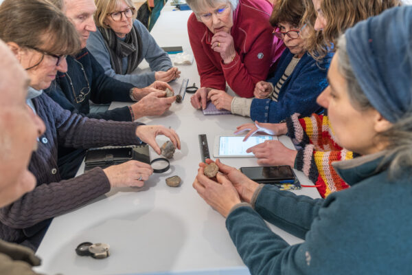 A group of people are recording sizes and shapes of different lithics found in the local area. They are sitting at a table with rulers, phones and magnifying glasses.