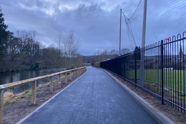 A new path can be seen in the middle of the picture. It is made of tarmac and very smooth. To the left of the path is a wooden fence and then the River Tweed. To the right, a high iron fence then the park green.