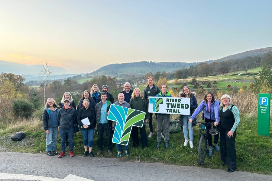 A group of people stand outside with a hilly landscape behind them. They are holding signs which have the River Tweed Trail logo on them and one person is on a bike.