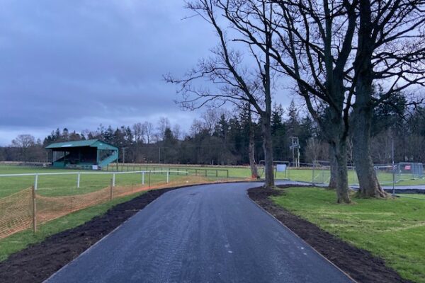 A new path is in the centre of the image. It is tarmac and edged with grass. Trees can be seen on the right side and on the far left a local football club stand.