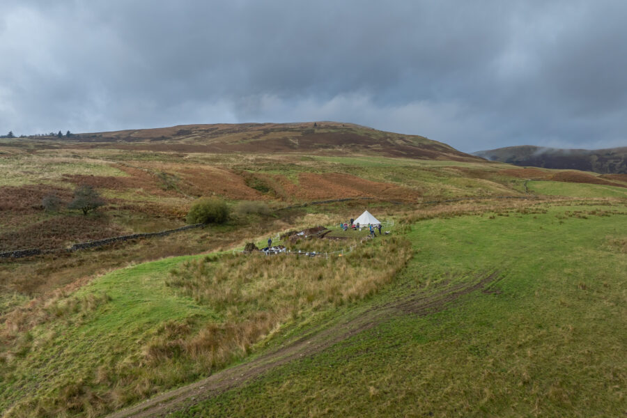 An aerial photograph of a a hilly landscape; a white bell tent sits on a hillside with an archaeological trench downslope from it