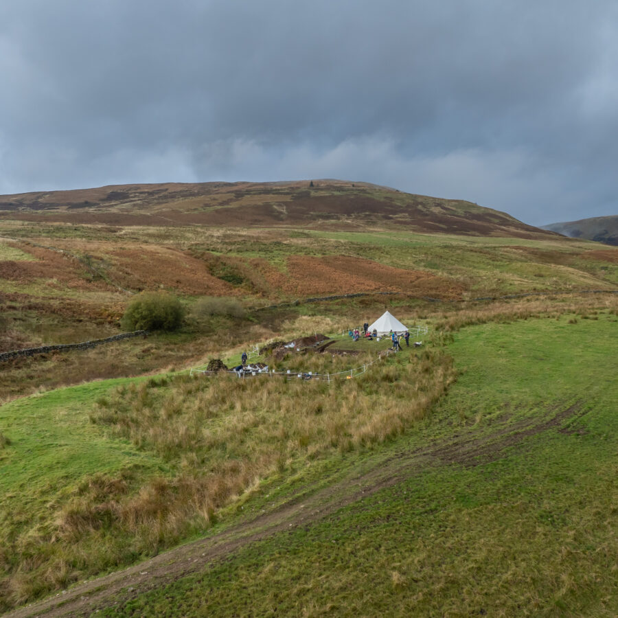 An aerial photograph of a a hilly landscape; a white bell tent sits on a hillside with an archaeological trench downslope from it