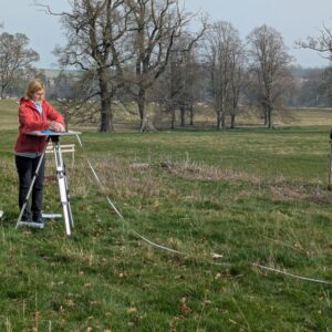 A woman wearing a red coat, standing in a grassy field at a tripod with a drawing board and alidade on the top, with a measuring tape dangling down the side