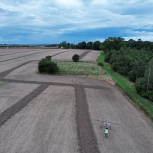 Aerial shot of an archaeologist pushing a wheeled geophysical survey instrument across a large arable field