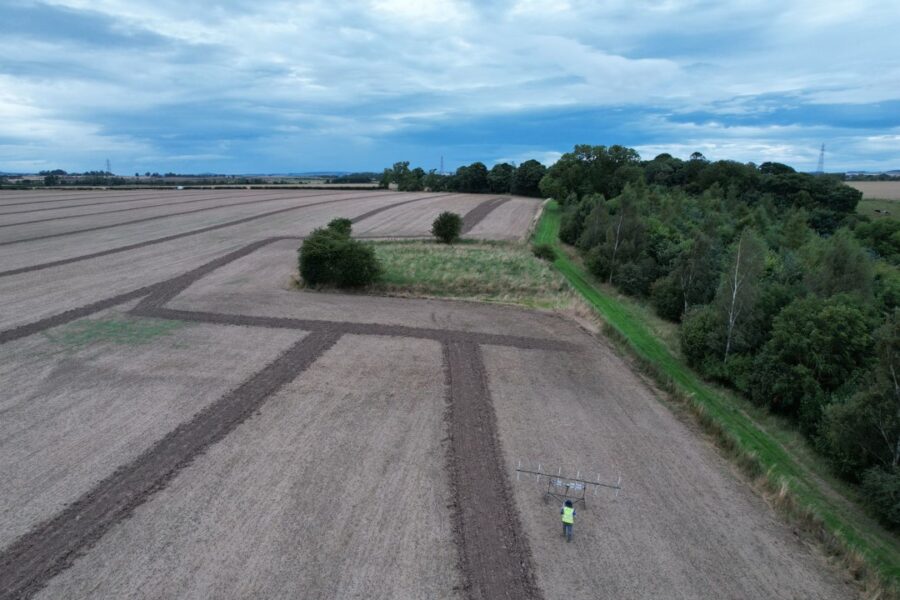 Aerial shot of an archaeologist pushing a wheeled geophysical survey instrument across a large arable field