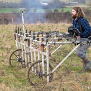 A man pushing a wheeled geophysical survey instrument across a tussocky field